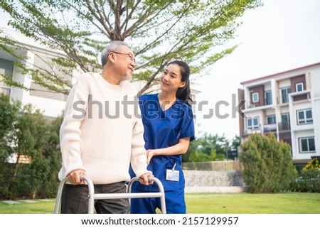 Asian young caregiver nurse support senior older male walking outdoors. Specialist girl doctor help and take care of elderly mature man patient doing physical therapy in public park at nursing home. Royalty-Free Stock Photo #2157129957