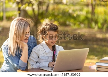 Mother helping her little daughter to use laptop computer. Child studying at home doing her homework or having online lesson. Homeschooling concept.