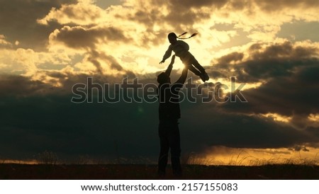 Dad plays with his daughter, throws child up into sky with his hands, happy kid. Father of daughters play together in park against backdrop of sun and clouds. Silhouette of happy family outdoors.