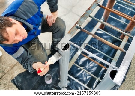Caucasian worker in overalls paints metal rack or ladder with paint brush. Real workflow in production hall. Industrial painter at work.