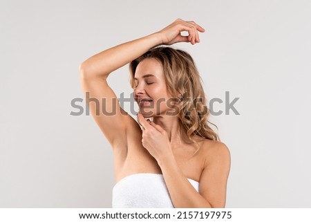 Beautiful caucasian woman smelling her fresh armpit while standing wrapped in towel over grey background, attractive millennial female raising hand up, enjoying underarm odor after bath routine
