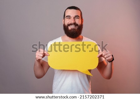 Smiling bearded man is holding a yellow bubble speech in front of his chest.