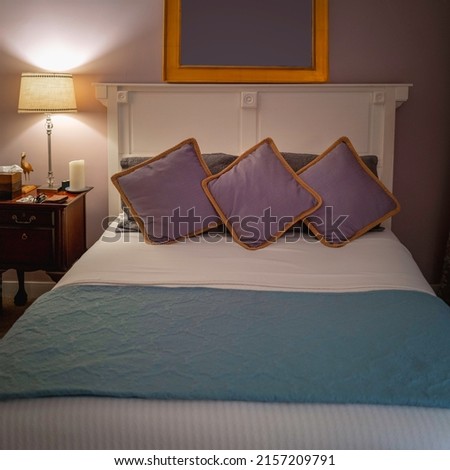 Vintage retro-style interior picture of bed with purple pillows, blue blanket, white linen sheet, night stand, and empty photo frame on the wall