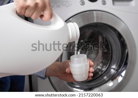 Against background of drum of steel-colored washing machine, woman pours liquid washing gel into plastic cap. A girl in a white T-shirt carefully pours a transparent conditioner for flattening laundry Royalty-Free Stock Photo #2157220063