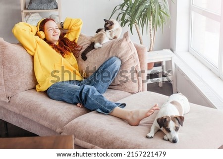 Young Woman listening music in headphones, Teen girl lying on sofa with cat and dog in living room at home
