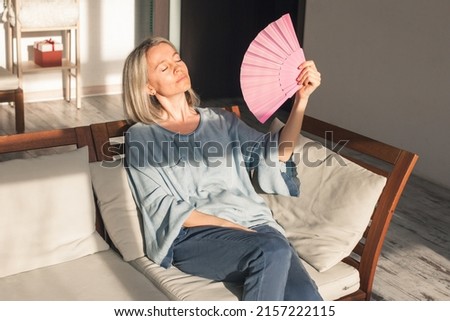 Side view sweaty middle aged lady using paper waving fan, suffering from high temperature inside. Unhappy middle aged woman feeling unwell, cooling herself at home with no air conditioning system. Royalty-Free Stock Photo #2157222115