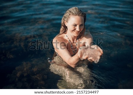 A blonde girl with long hair, in a striped swimsuit, swims in the ocean, laughs, holds a jellyfish in her hands on a bright sunny day.