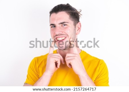 Happy Young caucasian man wearing yellow t-shirt over white background with toothy smile, keeps index fingers near mouth, fingers pointing and forcing cheerful smile