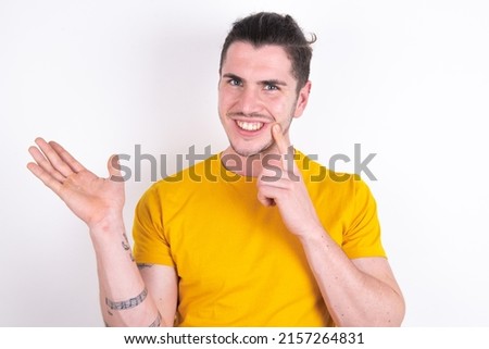 Positive Young caucasian man wearing yellow t-shirt over white background advert promo touch finger teeth