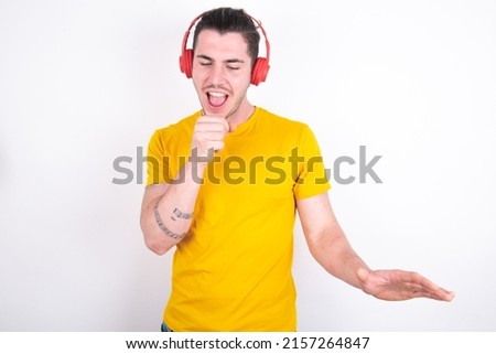 Happy Young caucasian man wearing yellow t-shirt over white background sings favourite song keeps hand near mouth as if microphone wears wireless headphones, listens music