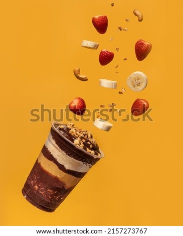 Acai cup, layers of granola, ground peanuts, and pieces of fruits and nuts flying in the air, yellow background. Royalty-Free Stock Photo #2157273767