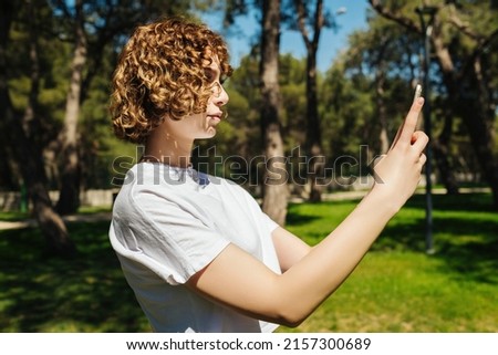 Young redhead woman wearing white tee taking smart phone by phone while standing on city park, outdoor.