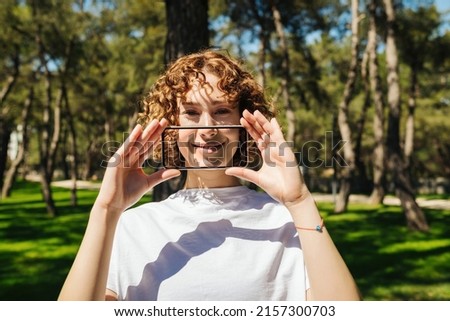 Portrait of a young redhead woman covering her mouth with transparent smartphone screen over green city park background, outdoor. Smiling on phone, digital and social media concepts.
