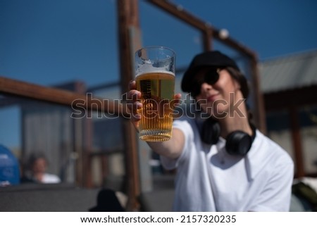 A young cheerful woman blurred in the picture, but her drink very sharp. She toasts with a glass of beer. Young stylish lady is holding a glass of beer in her hand. Cheers. 