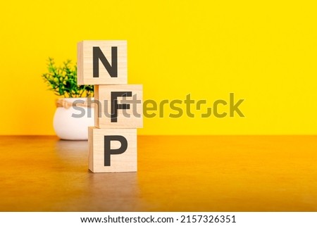 three wooden cubes with letters - nfp on yellow table, space for text in right. front view concepts, flower in the background. nfp - short for Non-Farm Payroll