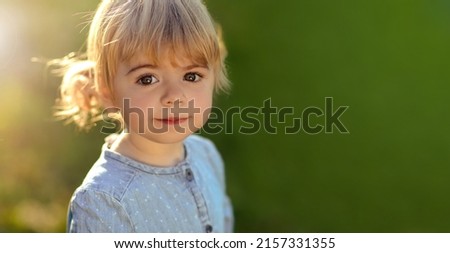 Close up of small blonde girl in blue jeans dress, with sunset rays in hair, smile, look at camera on green grass background. sunshine weather evening. Copy space for text, design. Extra wide banner