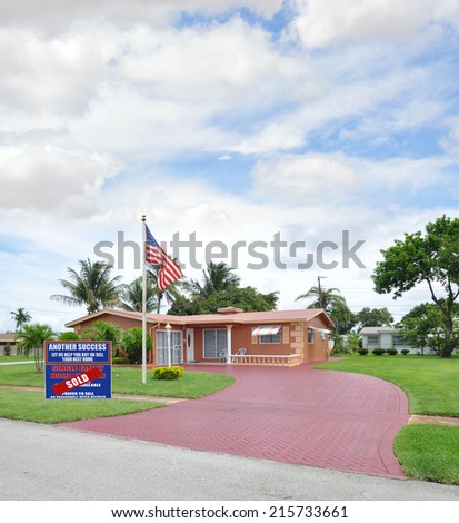 American Flag Pole Real Estate Sold (Another Success let us help you buy sell your next home) sign on front yard lawn of Suburban ranch style home residential neighborhood USA blue sky clouds