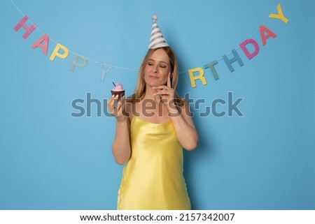 Pensive woman celebrating her birthday with cake in hands, wearing festive hat, enjoying surprise from friends, isolated on blue background