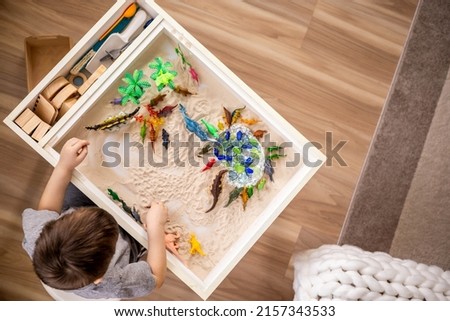 Top view cute little baby boy playing sensory box dinosaur world with kinetic sand table enjoy happy childhood. Male kid use carnivorous and herbivorous dinosaurs early development Montessori material Royalty-Free Stock Photo #2157343533