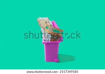 A pink garbage can full of different paper banknotes against green background. Surreal creative concept for waste of money editorial or banner. Symbolic design for economy inflation trend