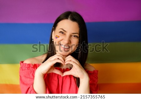 A young beautiful woman on the background of a rainbow LGBT flag folded her hands in the shape of a heart. A laughing person with a picture on his cheek.