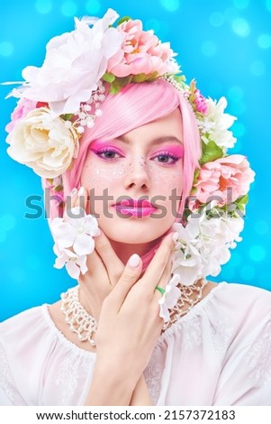 Gorgeous girl with bright pink make-up poses in colored pink wig and flower wreath on head. Blue studio background with lights. Beauty, makeup and hairstyle. 