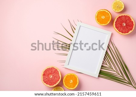 Summer weekend concept. Top view photo of white photo frame and ripe tropical fruits cut lemon orange grapefruit and palm leaves on isolated pastel pink background with empty space