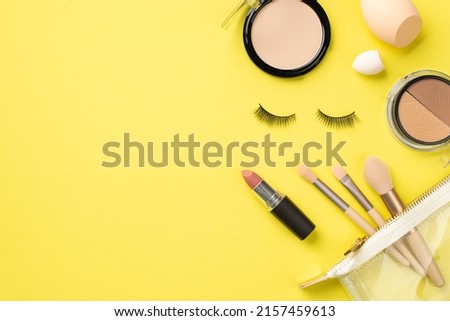 Make-up concept. Top view photo of compact powder contouring palette beauty blenders makeup brushes in cosmetic bag lipstick and false eyelashes on isolated yellow background