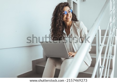 Beautiful business woman sitting on the stairs on stairs and working with laptop on the legs. Freelance girl have video call and look at laptop.
