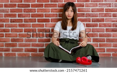 Full length of young attractive Asian woman in white t-shirt and green skirt sitting on the floor holding book with red headphone on the floor against orange brick wall background.