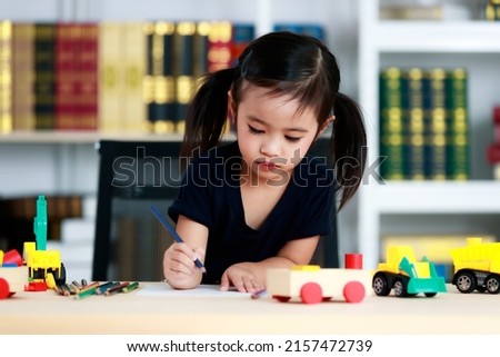 Small pretty pigtails hair preschooler kindergarten happy girl sit on chair drawing cartoon with colored pencils on paper on table full of plastic truck toys in living room at home in front bookshelf.