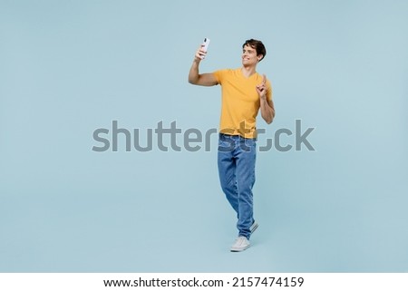 Full body young man wear yellow t-shirt doing selfie shot on mobile cell phone post photo on social network show v-sign isolated on plain pastel light blue background studio. People lifestyle concept Royalty-Free Stock Photo #2157474159