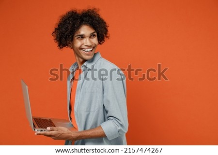 Young side view black man 50s in blue shirt t-shirt hold use work on laptop pc computer browsing look aside on copy space isolated on plain orange background studio portrait. People lifestyle concept
