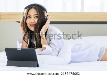Asian young pretty happy female teenager model in casual outfit wearing stereo headphone laying down smiling singing on bed in bedroom listening to song music playlist streaming from tablet computer.