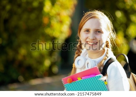 Portrait of smiling trendy girl in white sweatshirt with workbook, backpack and headphones near green school outdoors in the city.