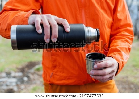A tourist man seems to be holding a thermos with hot tea in his hands, a man pours coffee into a mug in nature, tourist equipment, hiking equipment, a vacuum bottle. High quality photo