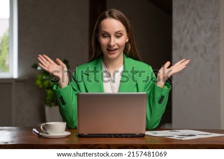 Cheerful woman gets happy looking at screen of laptop at wooden desk. Satisfied lady in green jacket sits in office rejoicing for deal with client