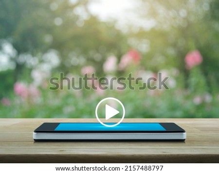 Play button flat icon on modern smart mobile phone screen on wooden table over blur pink flower and tree in park, Business music online concept