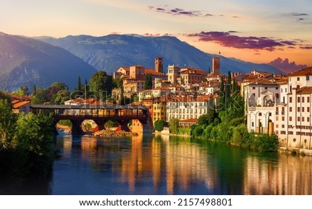 Bassano del Grappa, Veneto, Italy. Bridge Ponte degli Alpini at river Brenta. Panoramic view at old town with vintage building tower, wooden bridge at Alpine mountains scenic sunset landscape. Royalty-Free Stock Photo #2157498801