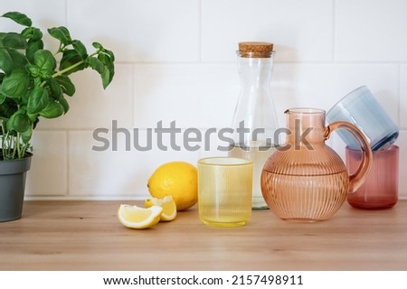 Colored glassware for drinks. Bottle, jug and glass for lemonade or water stand on wooden countertop or kitchen table, close to lemon and basil. Concept of antioxidant, detox and refreshment beverage Royalty-Free Stock Photo #2157498911