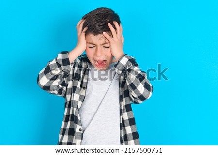Shocked panic caucasian kid boy wearing plaid shirt over blue background holding hands on head and screaming in despair and frustration. Royalty-Free Stock Photo #2157500751