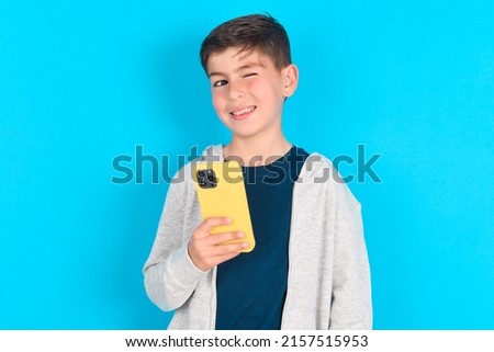 Pleased caucasian kid boy wearing grey hoodie over blue background using self phone and looking and winking at the camera. Flirt and coquettish concept.