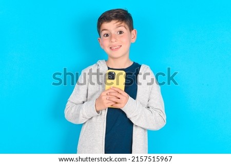 Smiling caucasian kid boy wearing grey hoodie over blue background friendly and happily holding mobile phone taking selfie in mirror.