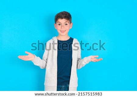 So what? Portrait of arrogant caucasian kid boy wearing grey hoodie over blue background shrugging hands sideways smiling gasping indifferent, telling something obvious. Royalty-Free Stock Photo #2157515983