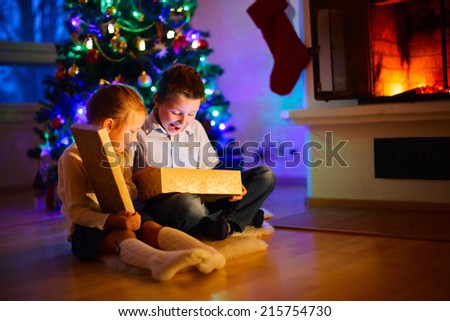 Little kids opening presents next to the tree and fireplace in a cozy home celebrating Christmas