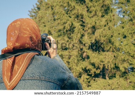 Elderly woman holds a camera in her hands against the background of a sunlight forest. Old lady takes aim at the viewfinder and takes a photo. Grandma photographs nature on a travel.