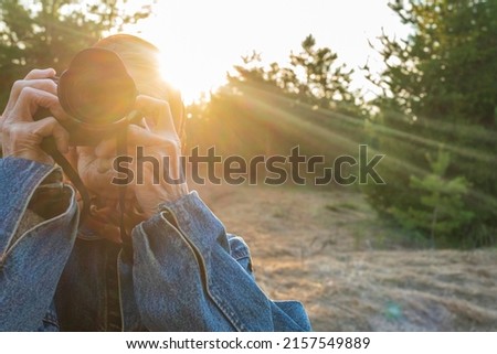Elderly woman holds a camera in her hands against the background of a sunset in the forest. Old lady takes aim at the viewfinder and takes a photo. Grandma photographs nature on a travel.