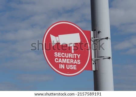 A red ‘Security Cameras in Use’ sign on the light pole with a tree and blue sky in the background.  