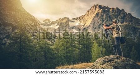 Panoramic Photo of Caucasian Photography Enthusiast in His 40s Taking Landscape Pictures Using Digital Camera with Tripod. Scenic Alpine Landscape.