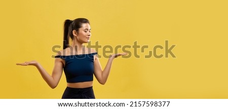 Smiling woman standing on knees, keeping hands palms up indoor. Portrait view of graceful girl in black top kneeling with arms bent at elbows, isolated on orange studio background. Concept of posing.